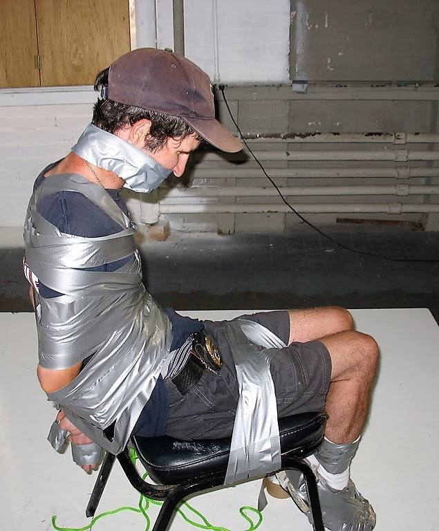 A man wearing a baseball cap and shorts is heavily restrained to a chair with layers of silver duct tape over his torso, neck, legs, and ankles.