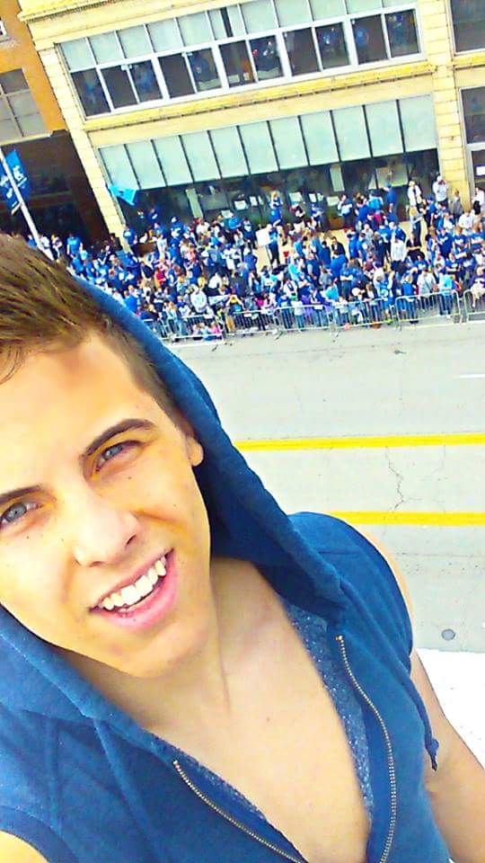 A smiling young man in a blue hooded sweatshirt takes a selfie at an outdoor public event.