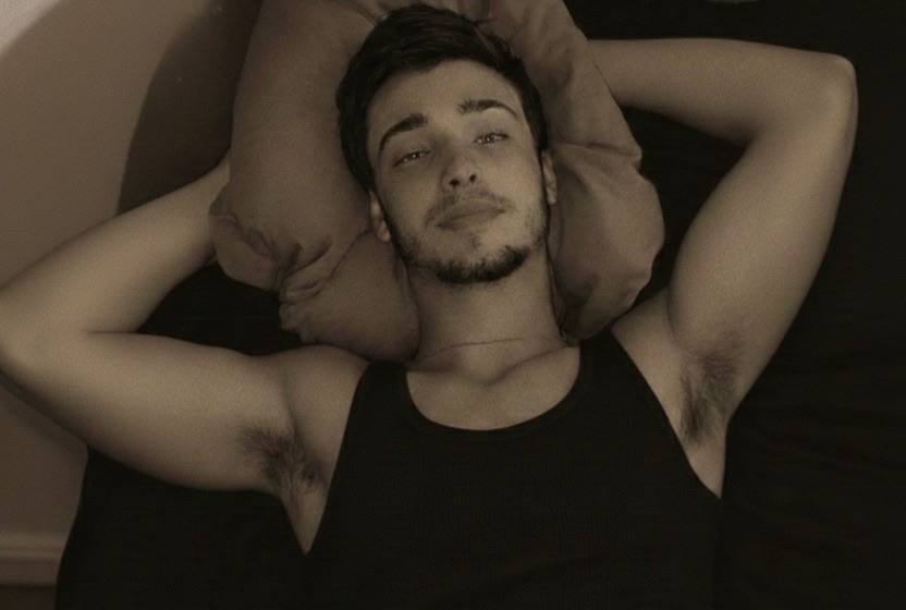 A young man with a trimmed beard and dark hair lies on a bed wearing a black ribbed tank top with his arms behind his head.