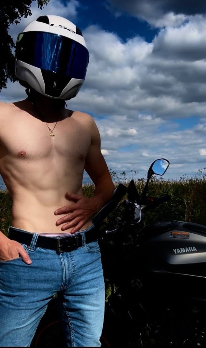 A shirtless man in a white motorcycle helmet and blue jeans poses outdoors next to a black Yamaha motorcycle.