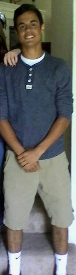 A young, lean man stands indoors wearing a blue henley, khaki shorts, and white Nike tube socks.