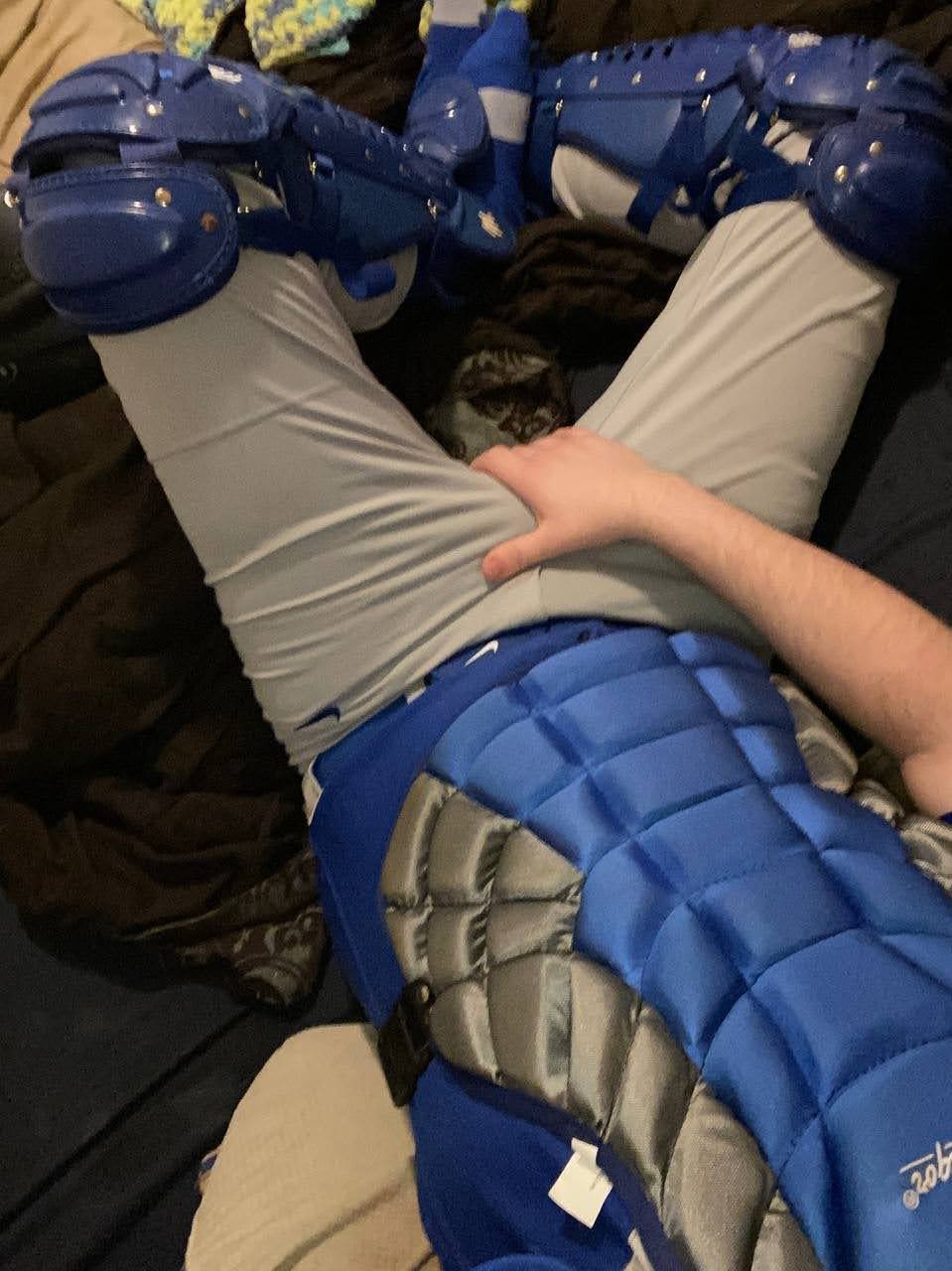 A person in full blue baseball catcher's gear and grey pants lies on a bed while grabbing their crotch.