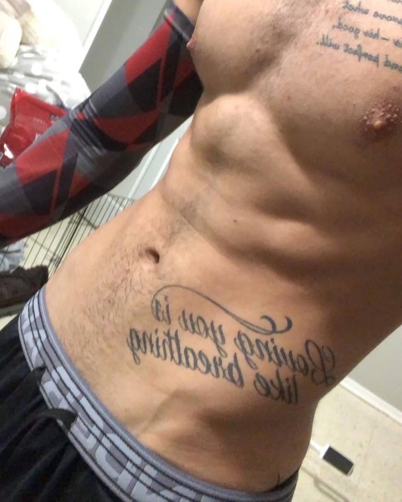 A muscular man with prominent chest and abdominal tattoos poses shirtless wearing a patterned compression sleeve and gym shorts.