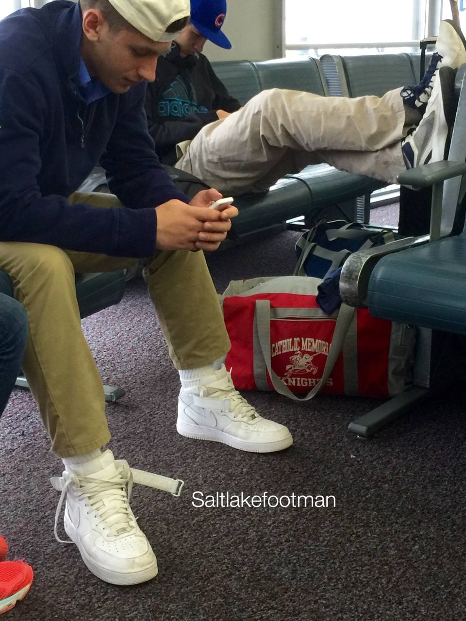 A young man sits in an airport terminal wearing white high-top sneakers and white tube socks while looking at his phone.