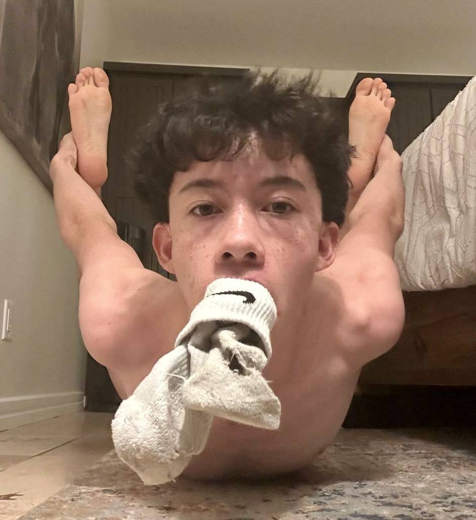A flexible twink lies on a rug, gagged with a white athletic sock and pulling his feet towards his head.