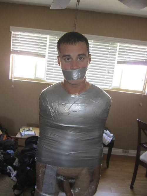 A man is bound around his torso and mouth with silver duct tape, with plastic wrap covering his lower body in a home setting.