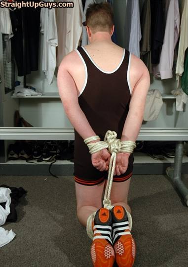 A man wearing a wrestling singlet kneels in a locker room with his wrists and ankles bound together behind his back with rope.