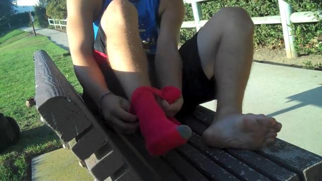 A young man in athletic wear sits on an outdoor park bench, focusing on his bare feet as he puts on red socks.