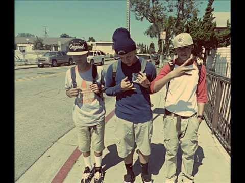 Three young men in casual streetwear, including baseball caps, beanies, and backpacks, pose on a sunny outdoor sidewalk.