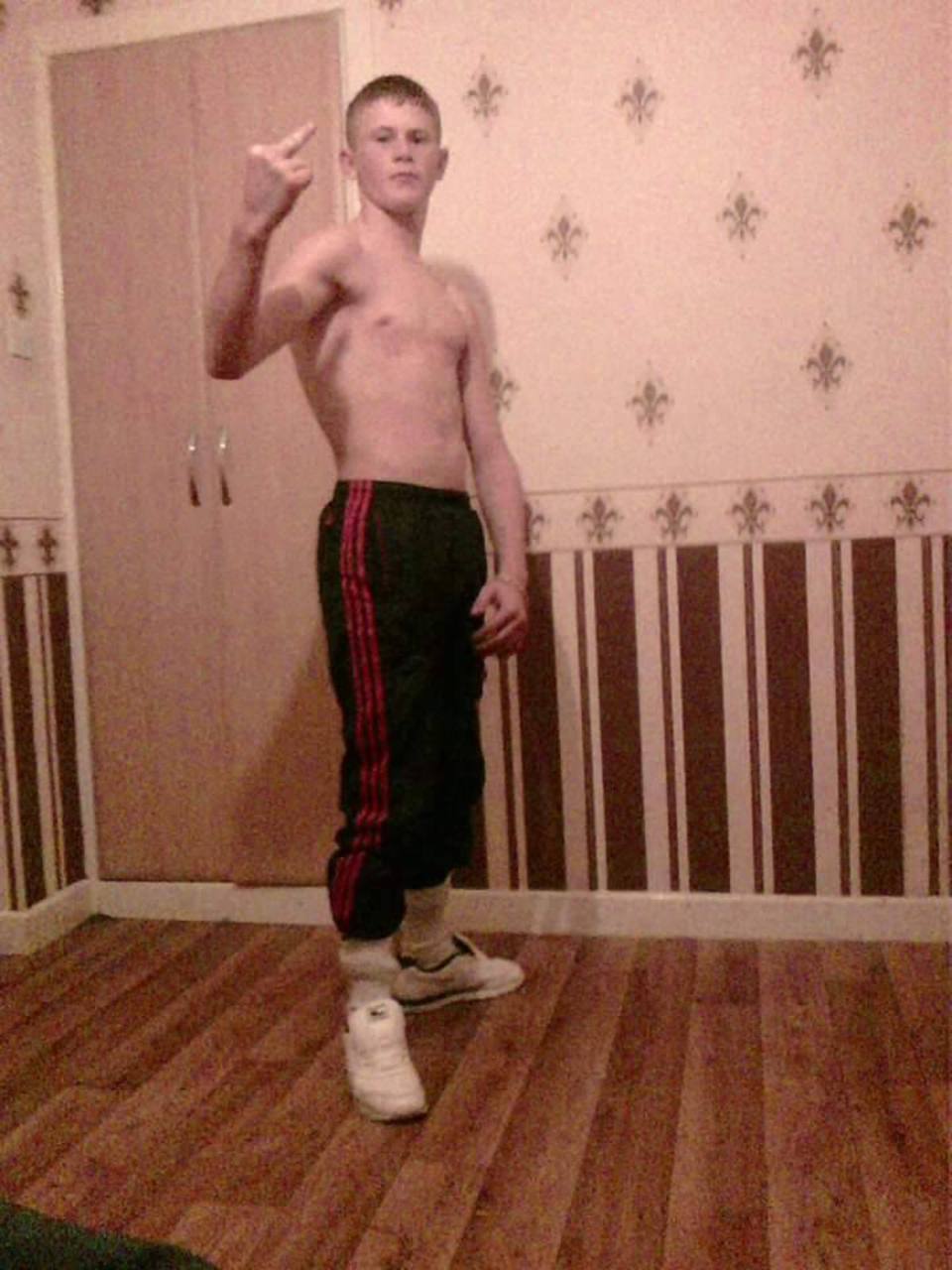 A shirtless young man with a buzzcut wears track pants tucked into socks and sneakers while posing indoors.