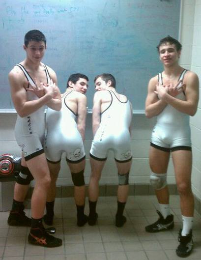 Four young men in white wrestling singlets and knee pads pose in a locker room, with two bending over to showcase their rear ends.
