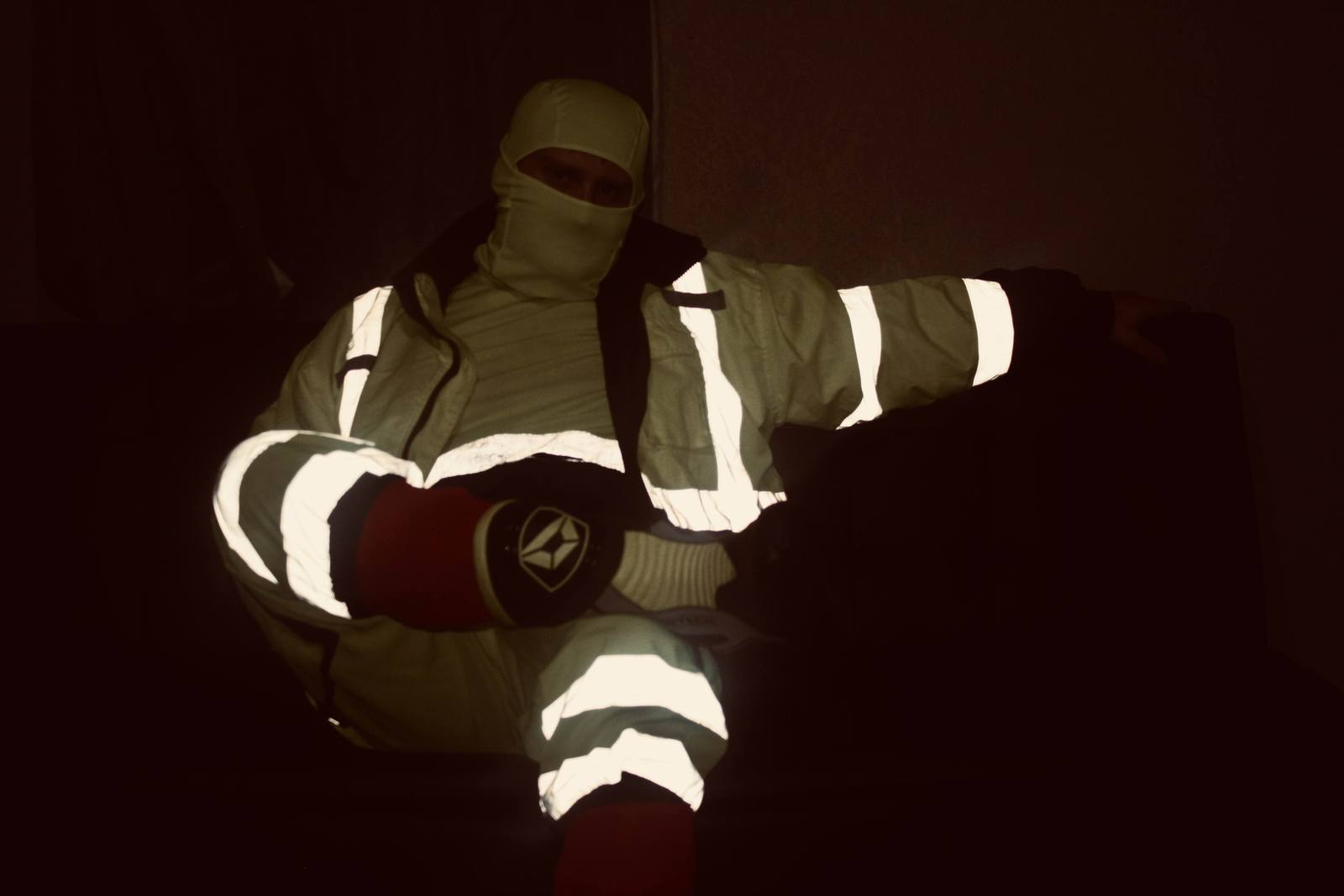 A man wearing high-visibility reflective workwear and a white balaclava sits in a dark room.