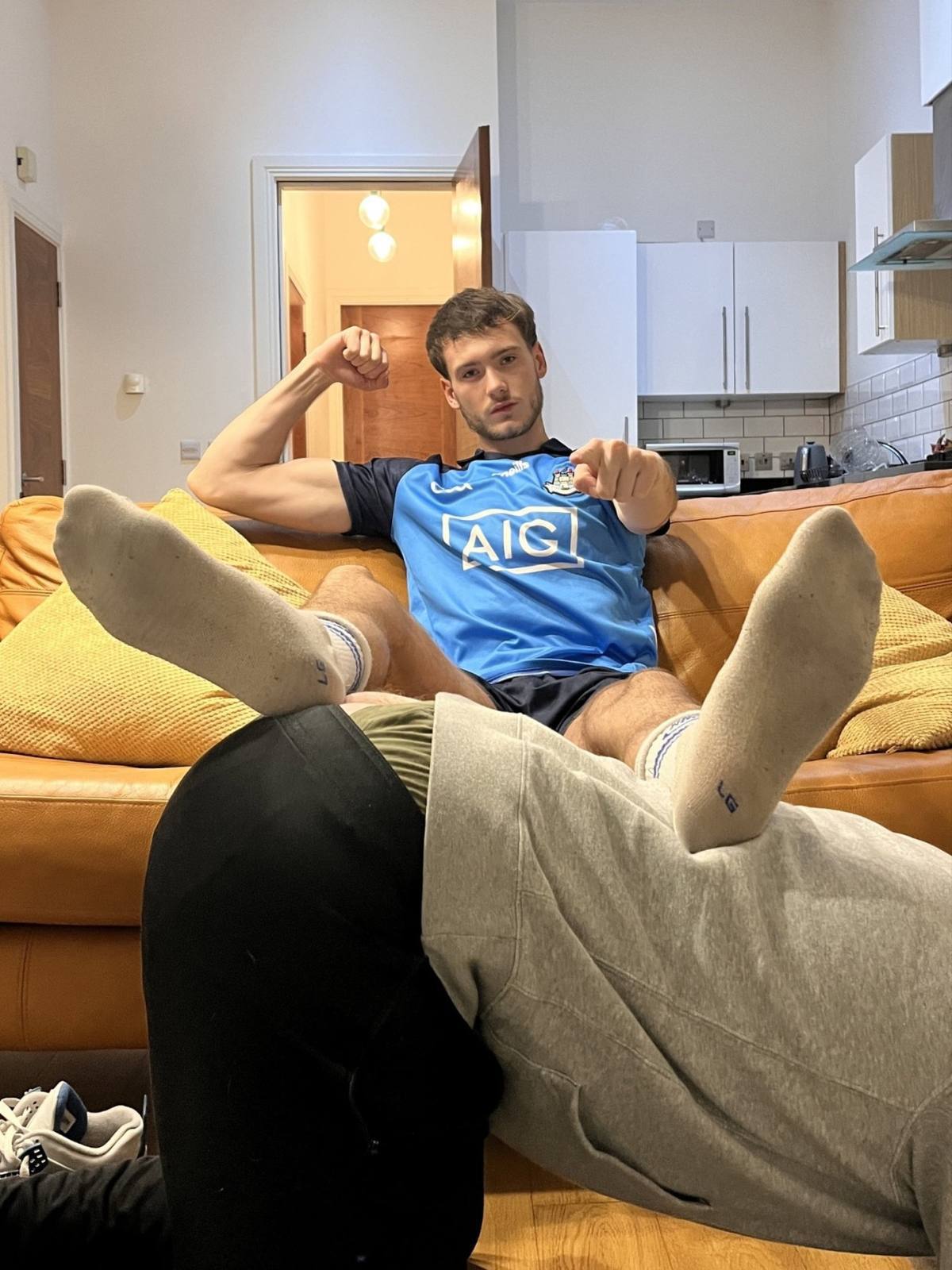 A muscular young man in a sports jersey flexes his bicep while resting his socked feet on a person kneeling in front of him.