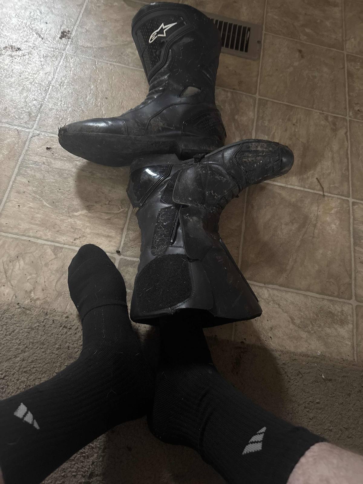 A person in black crew socks poses with a pair of well-worn Alpinestars motorcycle boots on a tiled floor.