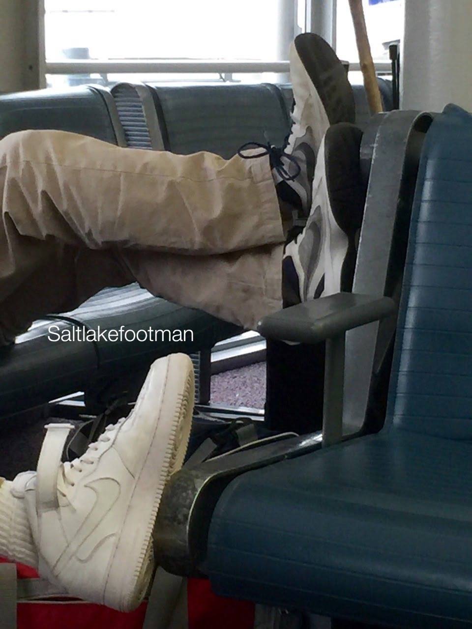 A man rests his feet on a terminal seat wearing white high-top sneakers and tube socks while another passenger rests their sneakers on a nearby armrest.