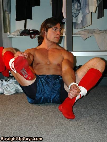 A shirtless athletic man sits on a locker room floor wearing blue nylon gym shorts and long red sports socks.