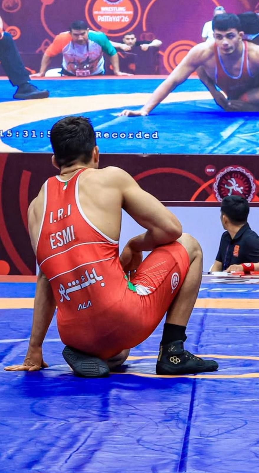 A muscular wrestler in a red lycra singlet kneels on a blue mat during a competition.