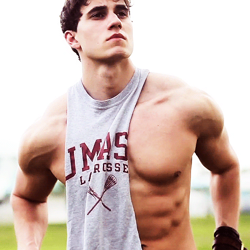 A muscular young man poses outdoors wearing a grey UMass lacrosse tank top pulled to one side to expose his defined chest and abdomen.