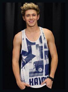 A young man with blonde hair smiles while wearing a sleeveless graphic tank top against a dark background.