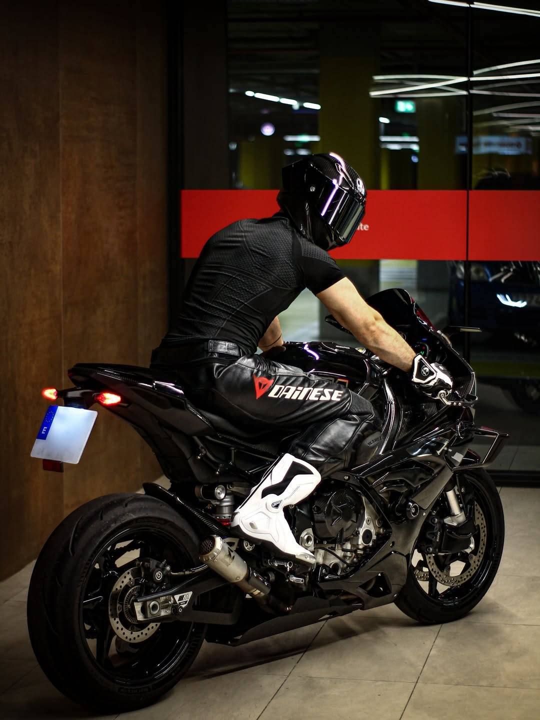 A muscular man wearing a helmet, compression shirt, and leather pants sits on a sport motorcycle in a garage.