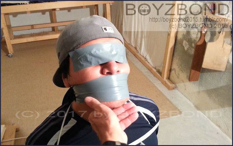 A young man is blindfolded and gagged with grey duct tape while an unseen hand tilts his chin up in a dungeon setting.