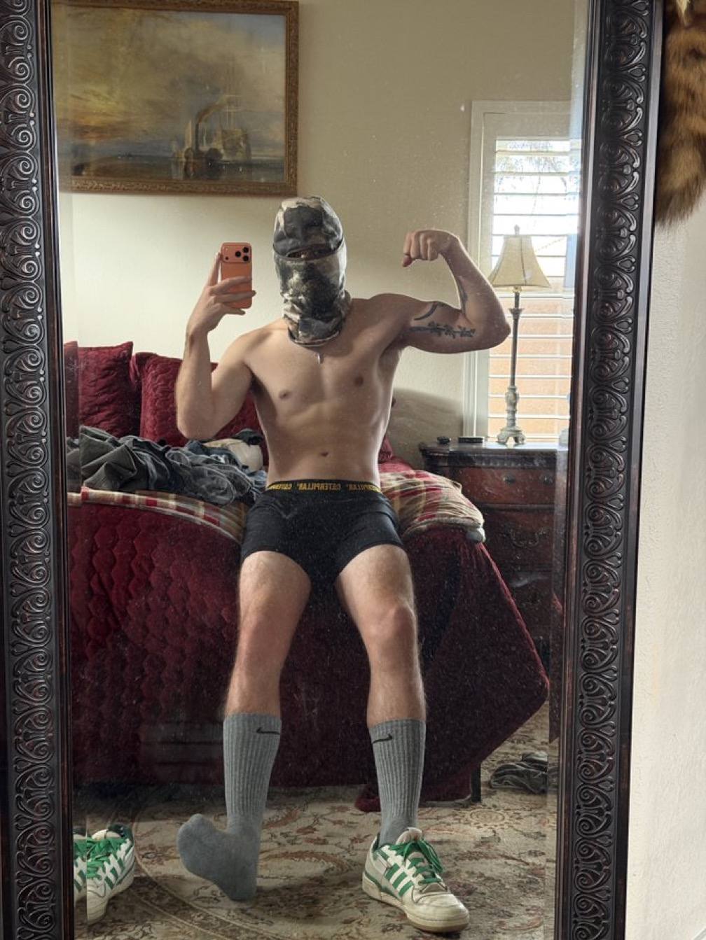 A muscular man takes a mirror selfie in a balaclava, underwear, socks, and sneakers.