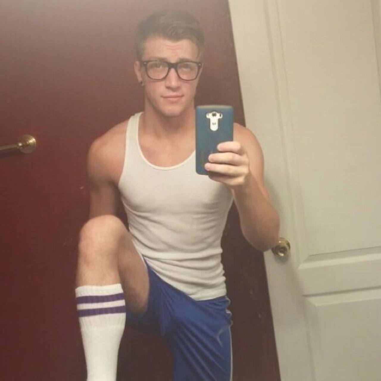 A young muscular man wearing a tank top, shorts, and tube socks takes a mirror selfie.