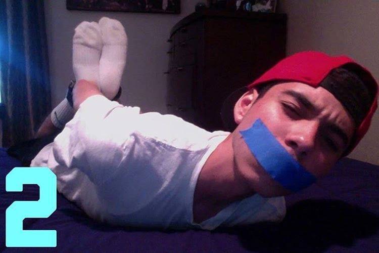A young man wearing a white t-shirt, red cap, and white socks lies on his stomach on a bed with blue tape covering his mouth.
