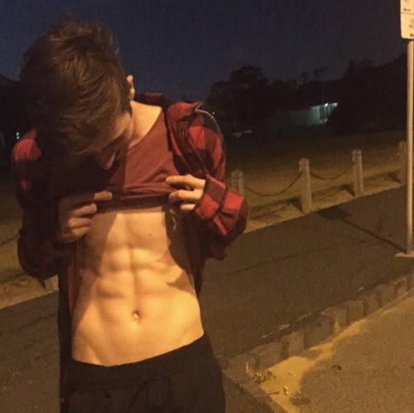 A lean young man pulls up his shirt to reveal his defined abdominal muscles outdoors at night.