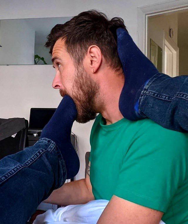 A bearded man in a green t-shirt submissively accepts a partner's blue-socked foot into his mouth while the other foot rests on his shoulder.