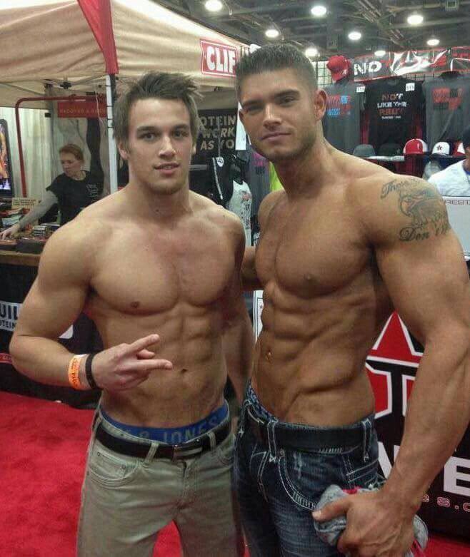 Two shirtless, muscular young men pose together at a fitness convention.