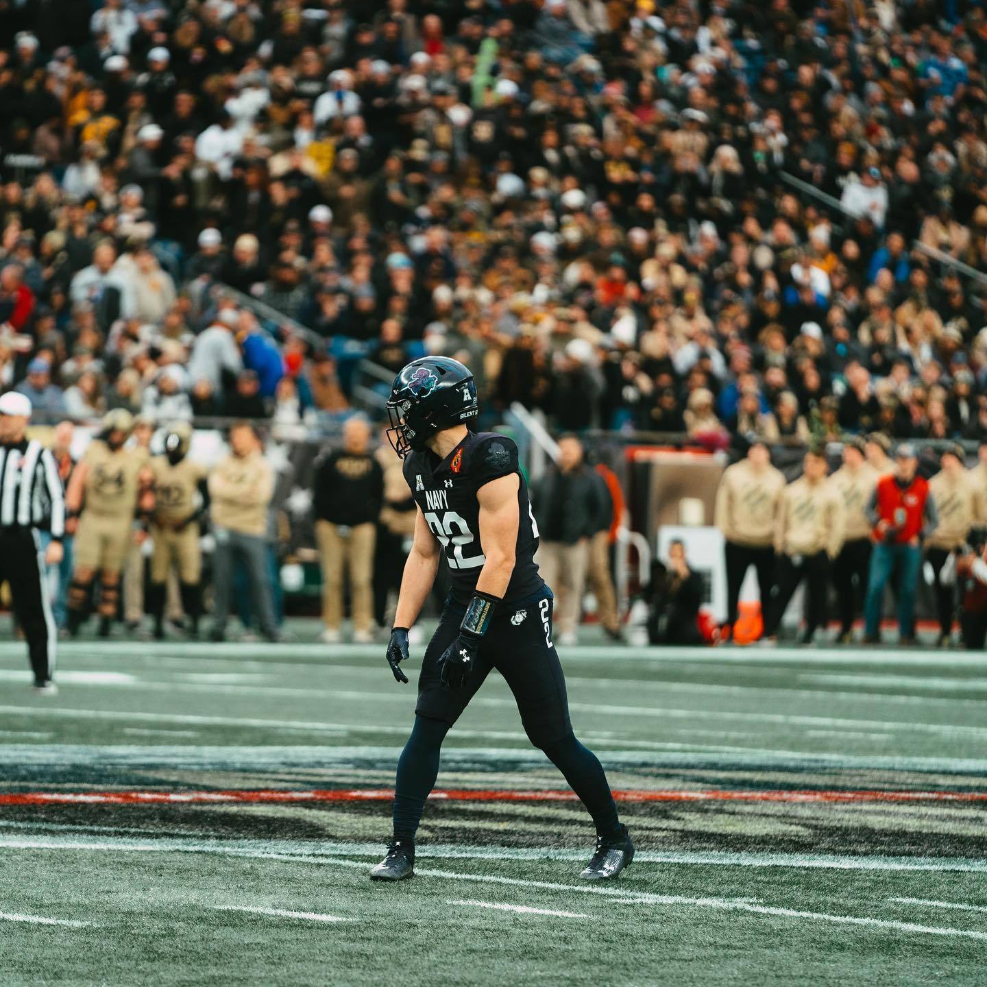A muscular athlete wearing a full Navy football uniform and helmet stands on the field during a game with a large crowd in the background.