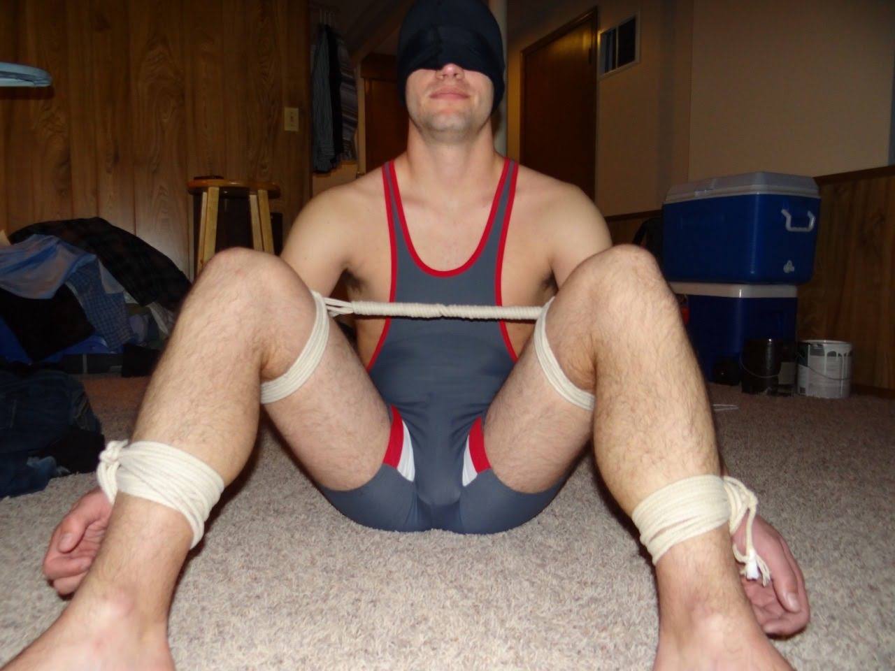 A blindfolded man in a grey and red wrestling singlet sits on the carpet with his legs restrained by white cotton rope.