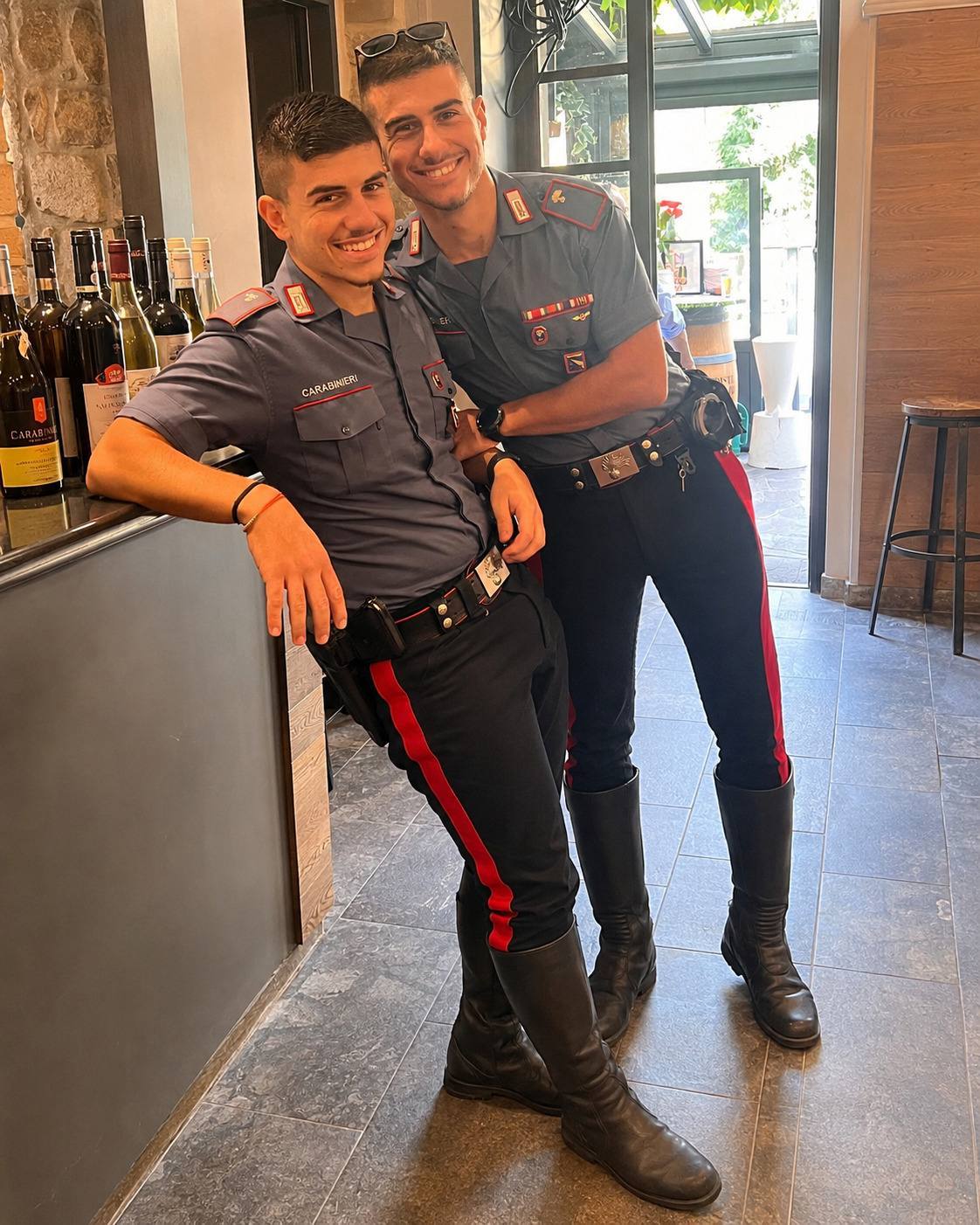Two fit men in Carabinieri uniforms, featuring tall black leather boots and utility belts, pose together in an indoor setting.