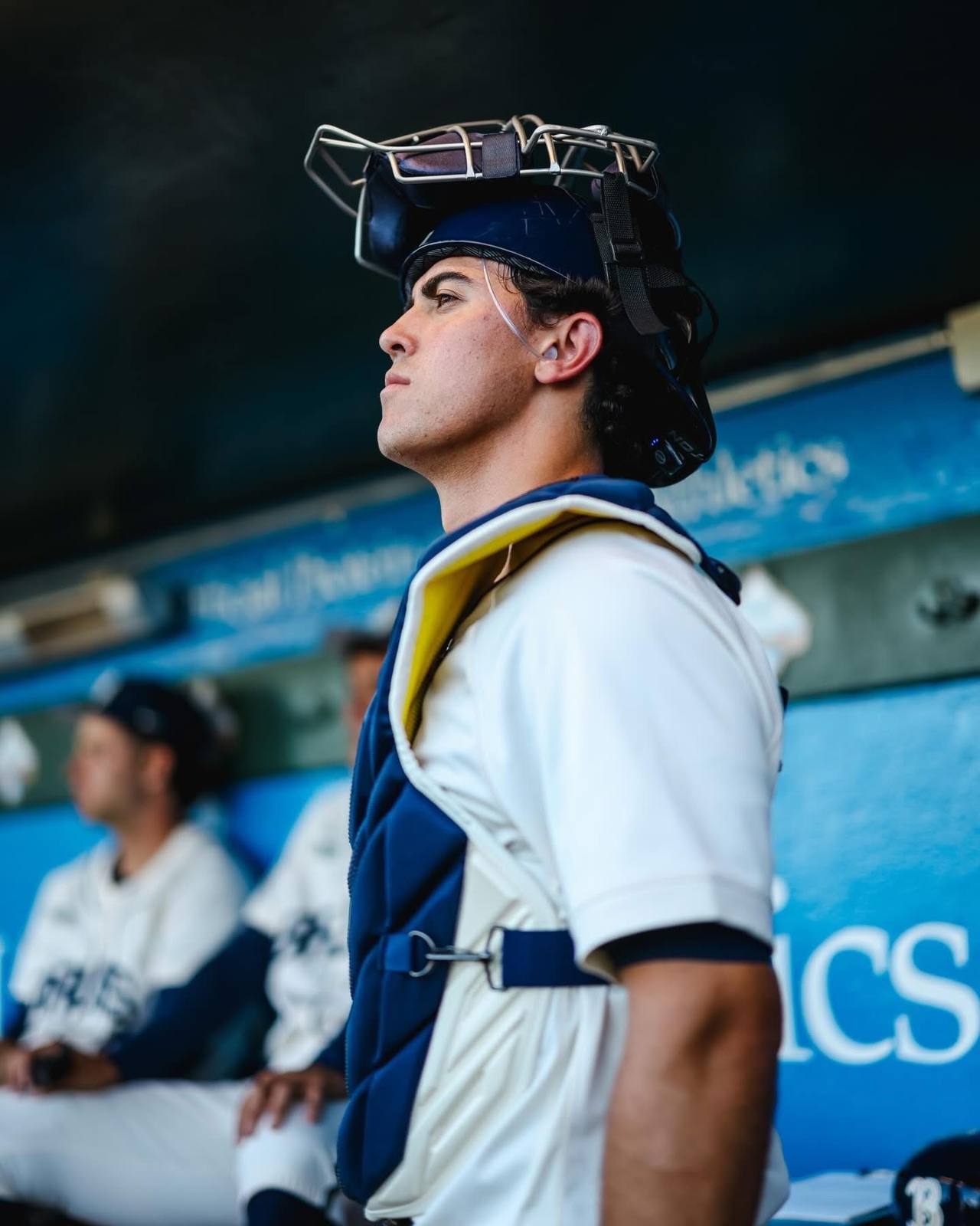 A baseball catcher in uniform with his mask pushed up stands in the dugout.