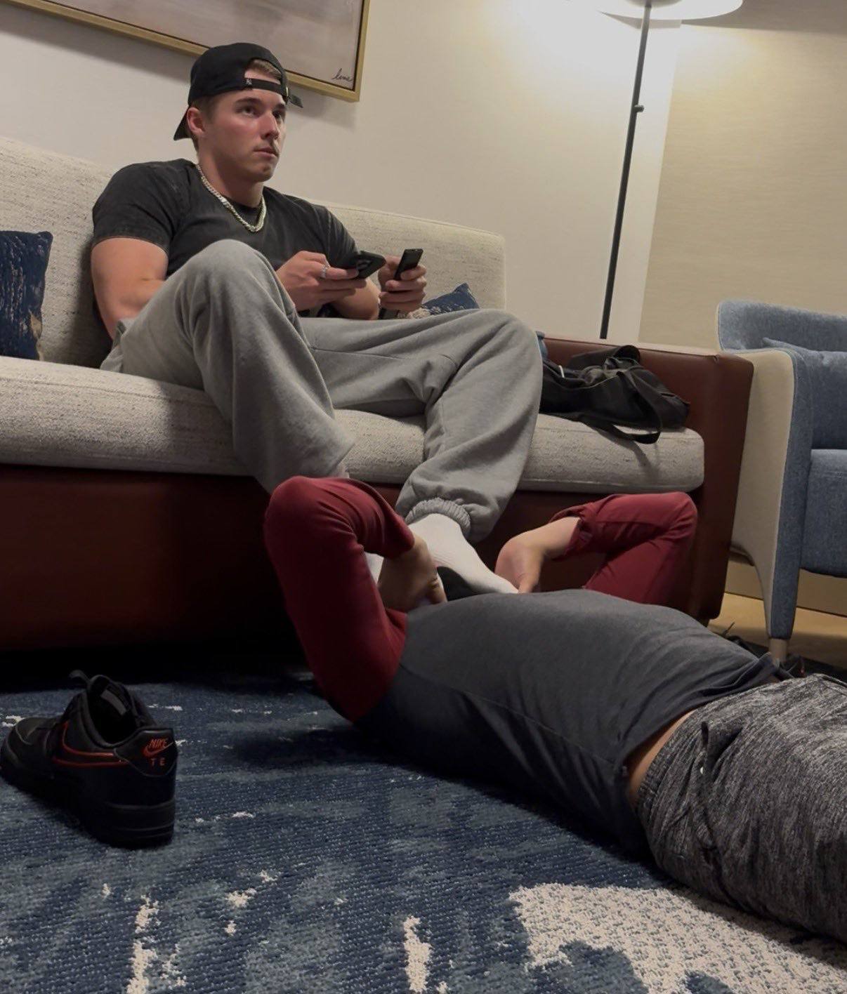 A muscular man relaxes on a couch while another man lies on the floor near his socked feet.