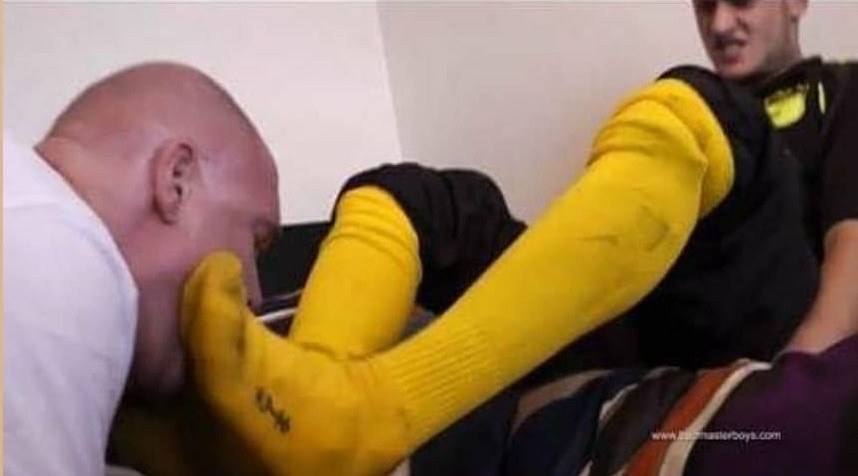 A man in a white shirt kisses the foot of another man wearing yellow athletic socks.