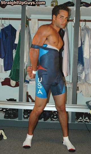 A muscular man in a USA wrestling singlet poses in a locker room with his arms pinned behind his back by the pulled-down straps.