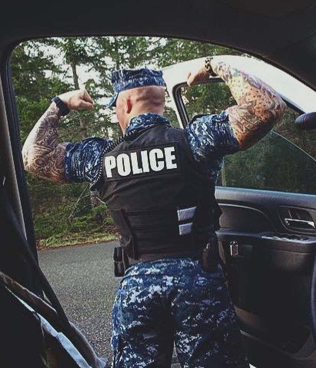 A muscular, tattooed man wearing a blue digital camouflage police uniform and tactical vest flexes his biceps while standing outdoors by a vehicle.