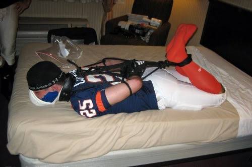 A bound male wearing a football jersey and orange socks lies hogtied face down on a bed.