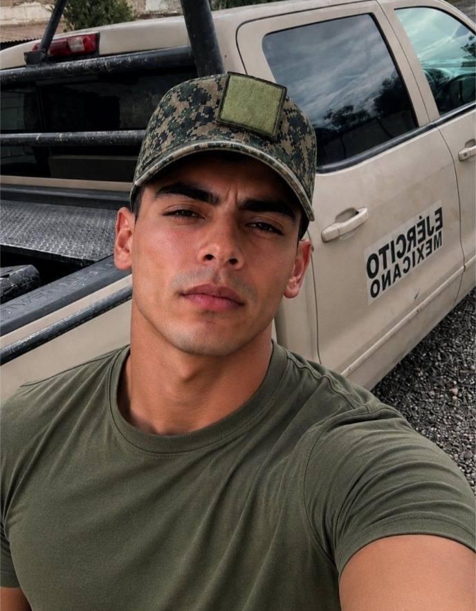 A muscular man in an olive green t-shirt and digital camouflage cap stands outdoors in front of a tan military truck.