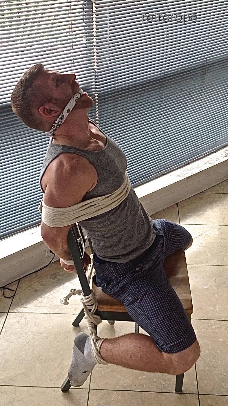 A muscular man wearing a grey tank top is bound tightly to a wooden chair with white rope and gagged with a bandana.