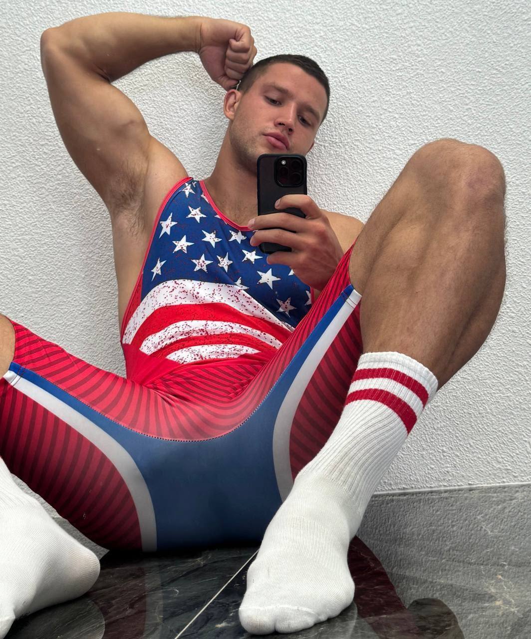 A muscular man with a buzzcut takes a mirror selfie while wearing a patriotic wrestling singlet and white tube socks.