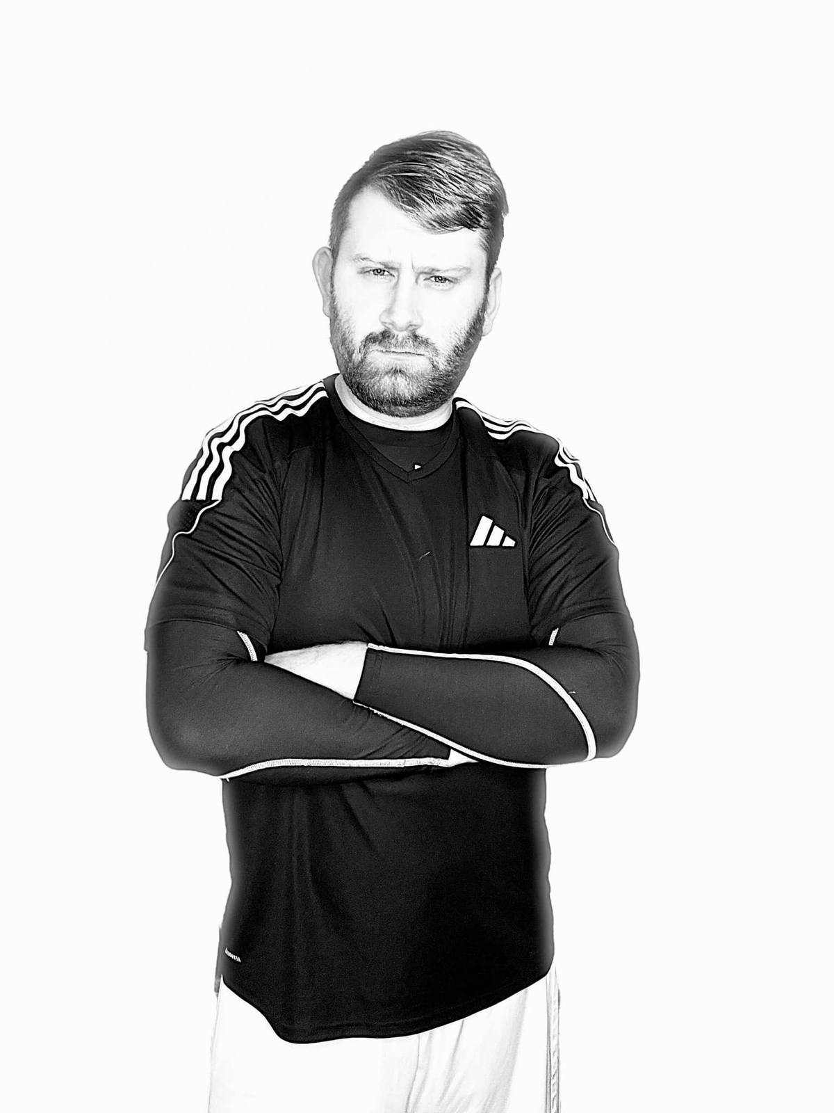 A bearded man wears a black Adidas jersey and compression sleeves with his arms crossed against a plain white background.