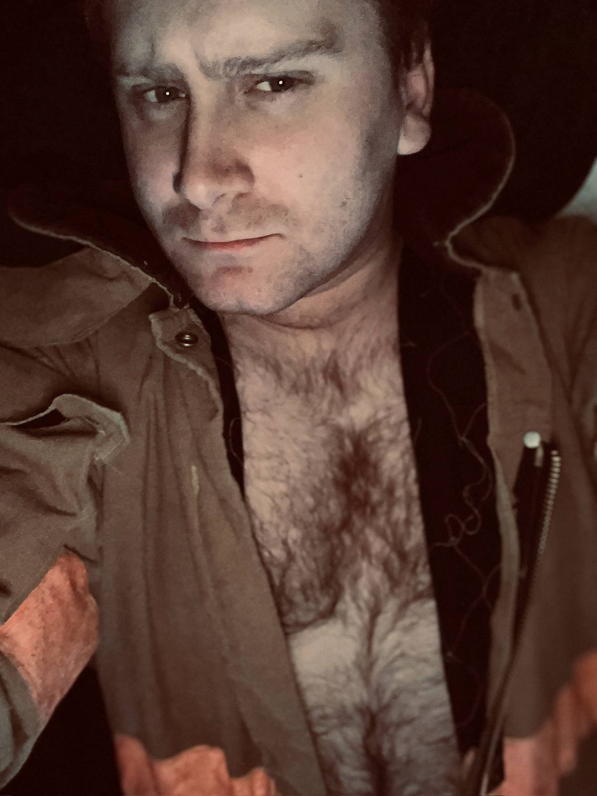 A man with a dense mat of chest hair poses in an unzipped tan flight suit.