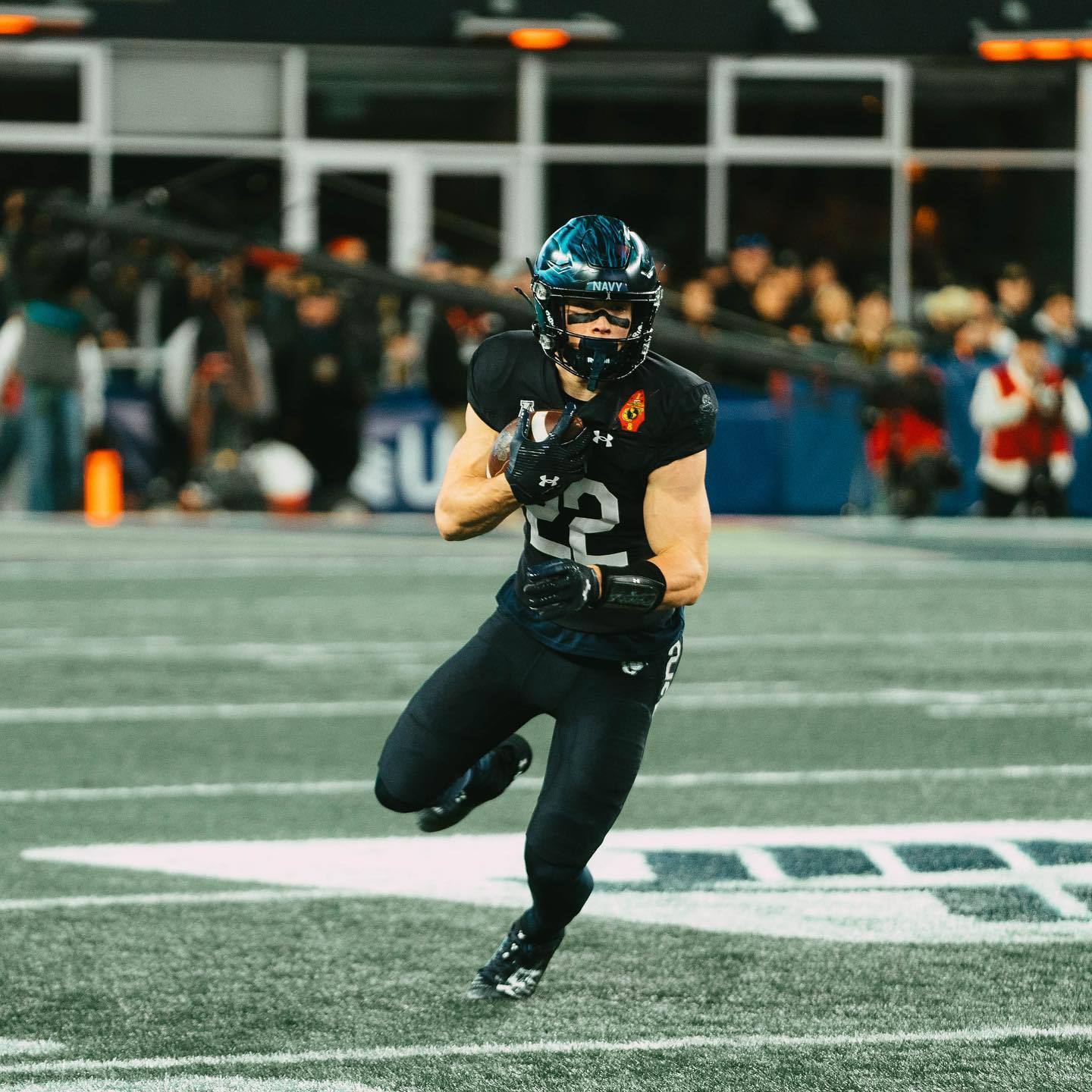 A muscular football player in a black uniform and helmet runs across the field carrying the ball.