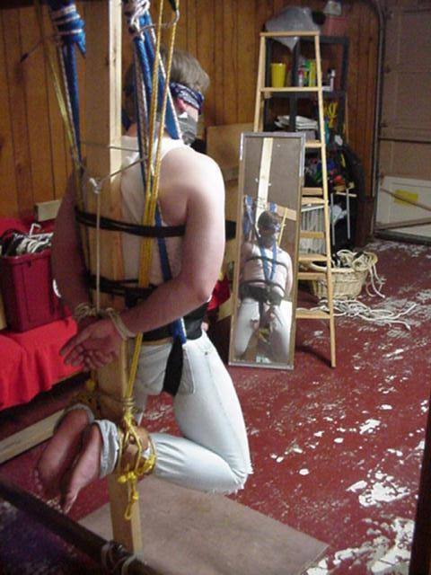 A man wearing denim jeans and a tank top is securely bound and suspended on a wooden post using ropes, straps, a blindfold, and duct tape gag.