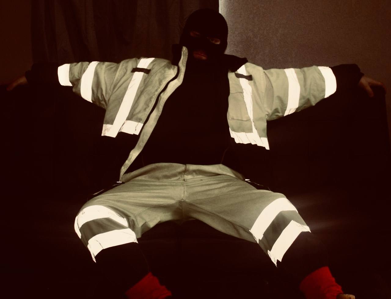 A person wearing high-visibility construction workwear and a black balaclava sits on a sofa in a dark room.