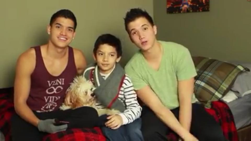 Two young men and a child sit together on a bed with a red plaid blanket, accompanied by a small white dog.