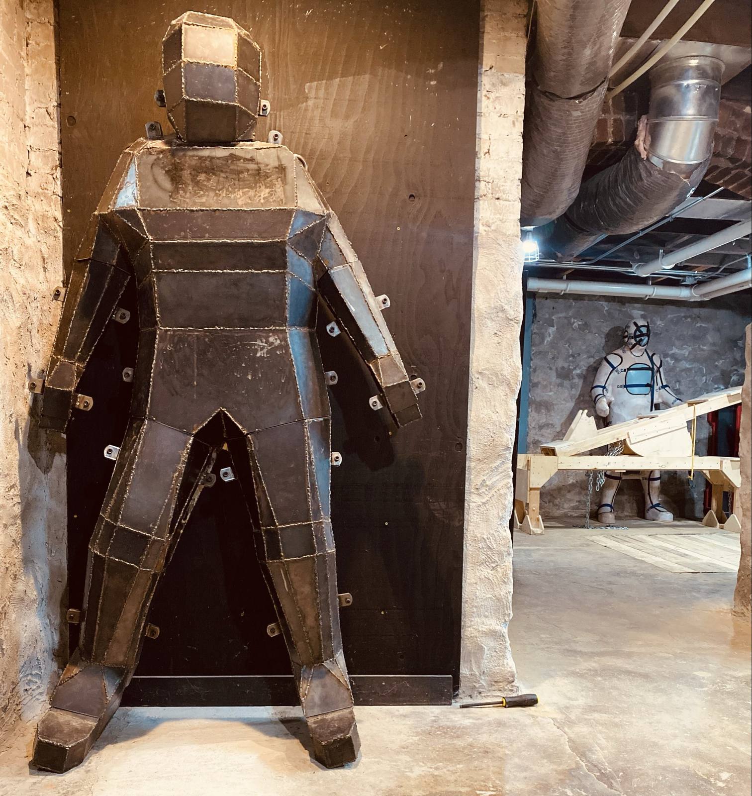 A large, geometric metal body-containment device stands in a dungeon while a figure in a white full-body suit and black harness stands in the background.